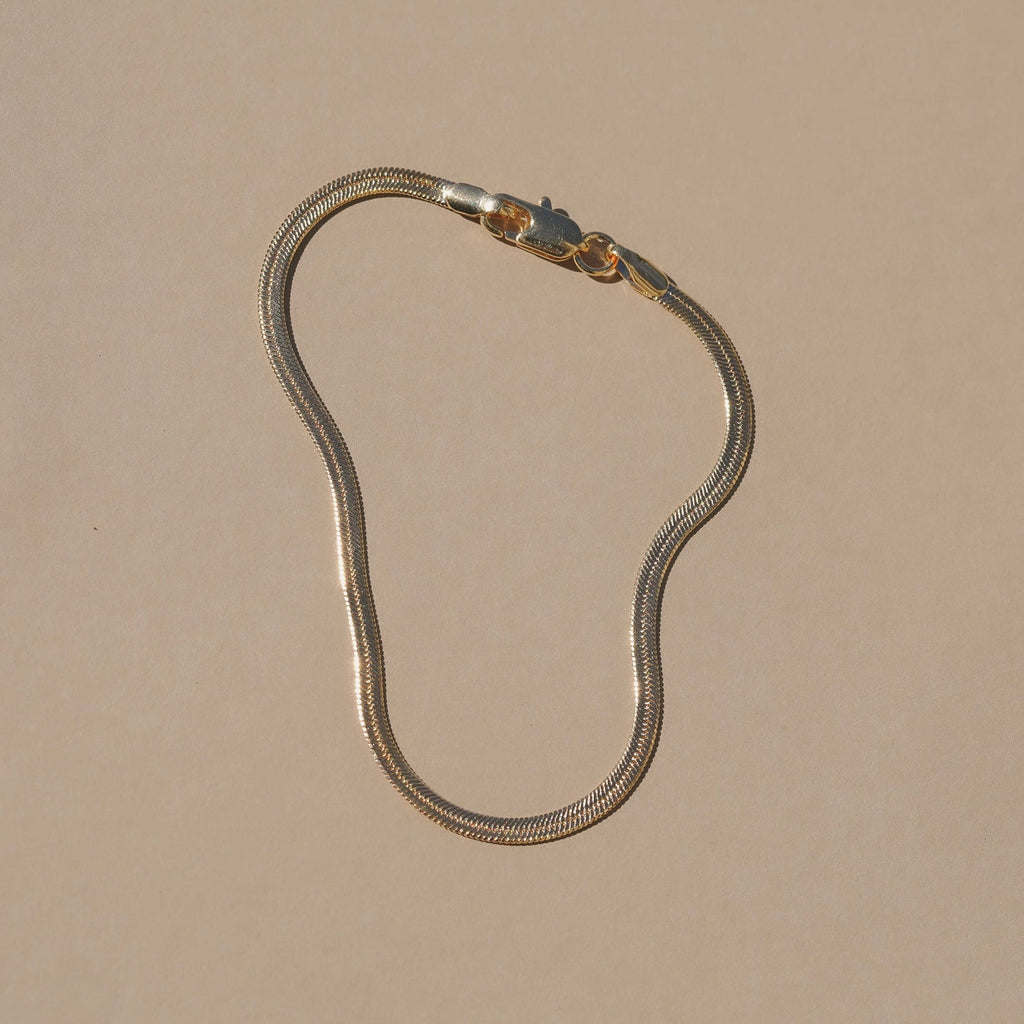 A gold-toned snake chain necklace with a lobster clasp, displayed on a solid beige background.