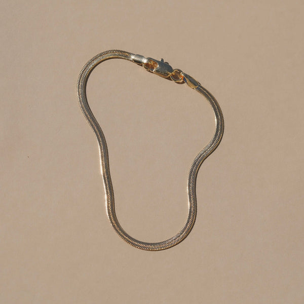 A gold-toned snake chain necklace with a lobster clasp, displayed on a solid beige background.
