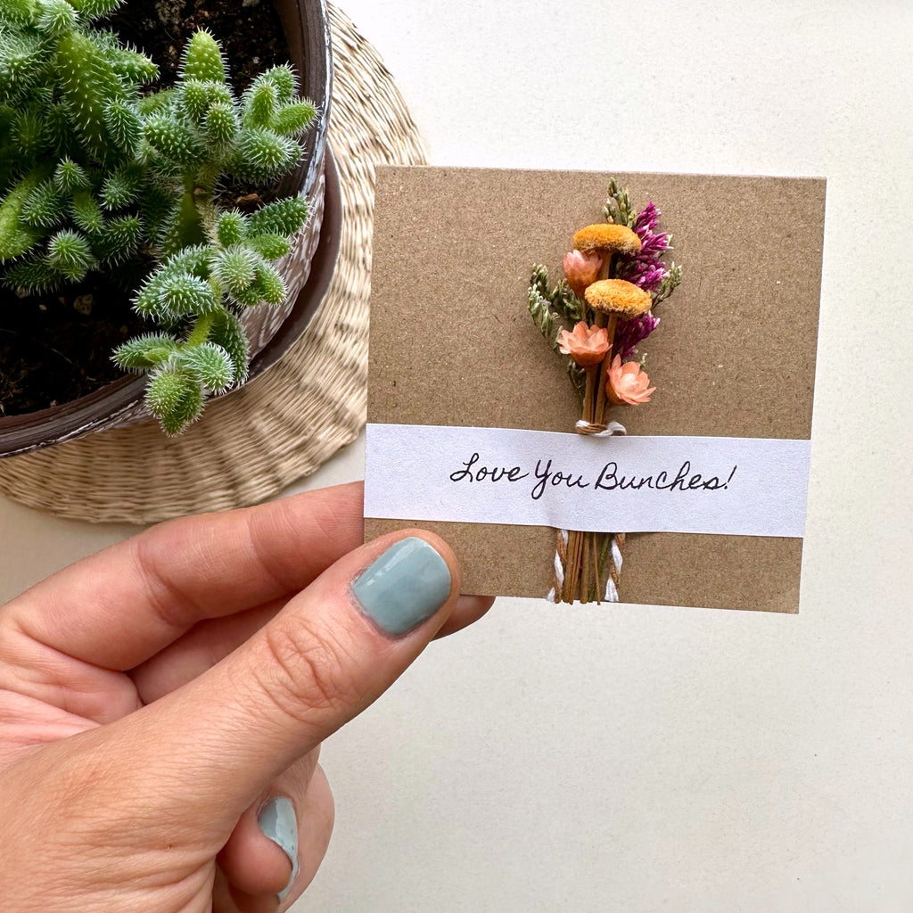 Hand holding a card with dried flowers and 'Love You Bunches!' text, next to a potted plant.