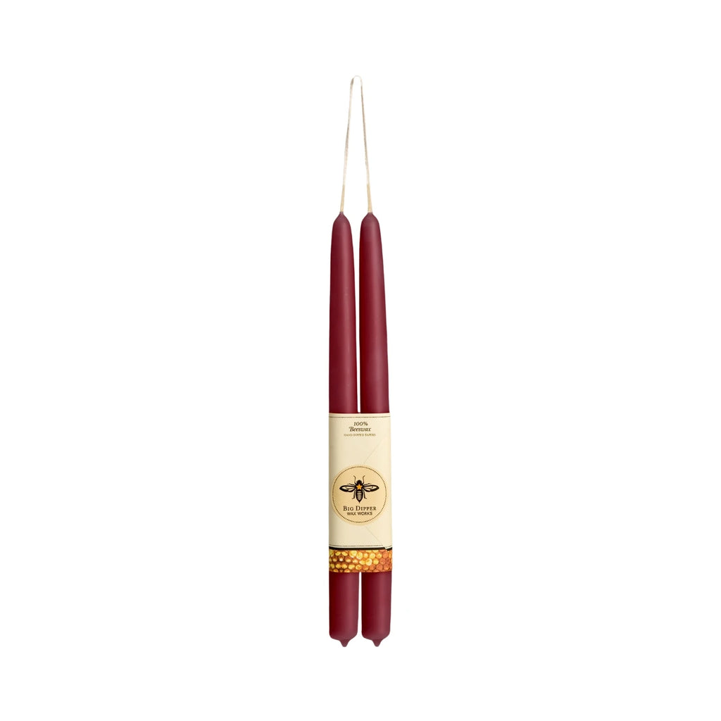 Two tall taper candles in deep burgundy red, wrapped together with a cream paper band featuring a gold bee logo and a gold patterned accent, suspended against a plain white background.