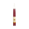 Two tall taper candles in deep burgundy red, wrapped together with a cream paper band featuring a gold bee logo and a gold patterned accent, suspended against a plain white background.