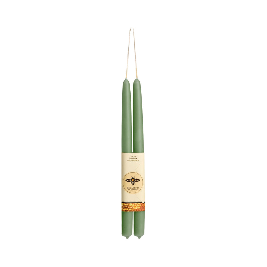 A pair of tall, slender sage green taper candles wrapped in a cream paper band with a bee logo, hanging against a solid white background.
