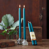 Three teal taper candles are displayed on a wooden surface, with two standing in clear glass holders and one wrapped in a cream paper band. The candles are lit, with flames visible, and are styled with rustic wooden boxes and a blurred green plant in the background.